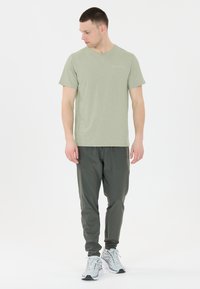 Man med ljusgrön kortärmad skjorta, mörkgrå joggerbyxor och vita sneakers, som står mot en vit bakgrund och tittar åt vänster.