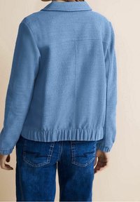 Jasnoniebieska polarowa kurtka z kołnierzem, teksturowana powierzchnia i elastyczny ściągacz u dołu. Połączona z dżinsami z dekoracyjnym przeszyciem na kieszeniach.