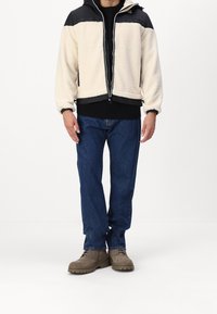 Fleece-takki, jossa on mustat nailonhartiat, vetoketju edessä ja elastiset hihansuut. Yhdistetty tummansinisiin farkkuihin ja ruskeisiin nahkakenkiin.