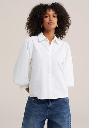 Femme aux cheveux bouclés portant des boucles d'oreilles créoles en or, un chemisier blanc à manches bouffantes et un jean bleu sur un fond neutre.