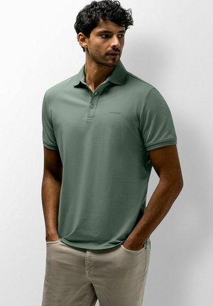 Man met krullend zwart haar, gekleed in een groen poloshirt en beige broek, staat met zijn handen in zijn zakken en kijkt opzij tegen een effen achtergrond.