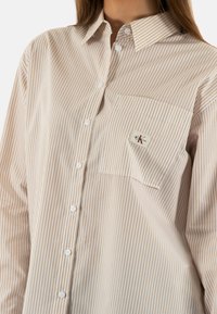 Camisa de rayas de color claro con rayas verticales en blanco y beige, con botones en la parte delantera, cuello y un bolsillo en el pecho izquierdo que presenta una etiqueta de logo.