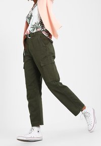 Olive green cargo pants with a relaxed fit, featuring multiple side pockets and a belt. Paired with a floral shirt and white sneakers.