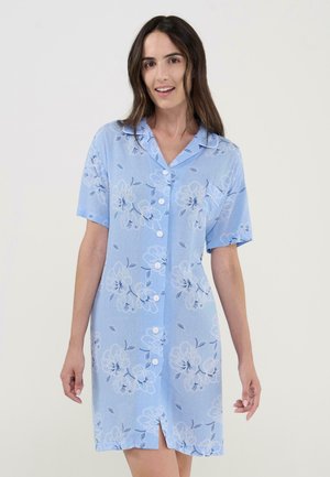 Mujer de cabello largo y oscuro, vestida con un vestido azul claro de manga corta con botones y estampado floral, sonriendo frente a un fondo liso.