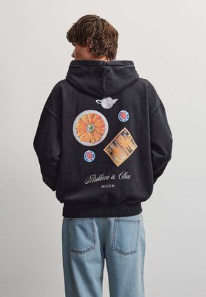Persona che indossa una felpa nera con cappuccio con grafica colorata sul retro di baklava, teiera, tazze e tavolo da backgammon, abbinata a jeans azzurri chiari.