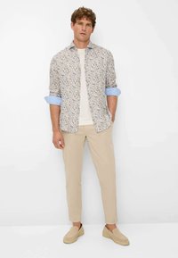 Leicht gemusterte Button-Up-Bluse mit hochgekrempelten Ärmeln, beige Chinos und braunen Slip-On-Schuhen. Die Bluse zeichnet sich durch florale Designs in gedämpften Farben aus.