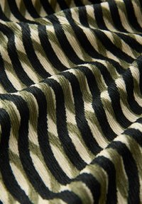 Tekstil yang menampilkan pola zigzag dalam warna hijau zaitun, krim, dan hitam. Kain memiliki permukaan bertekstur, beralur, yang menambah kedalaman visual.
