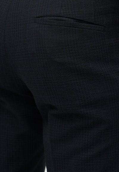 Κοντινό πλάνο σκούρου υφασμάτινου παντελονιού με διακριτικό καρό μοτίβο και λεπτομέρεια πίσω τσέπης με επένδυση.