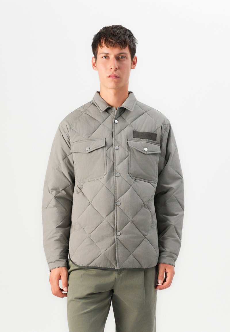 Young man wearing a gray quilted jacket with buttoned chest pockets and matching gray pants, standing against a white background.