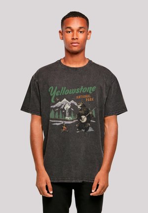 Young man wearing a black Yellowstone National Park t-shirt with a bear playing a banjo and a campfire in front of mountains and trees.