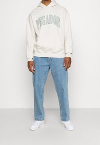 Felpa beige con testo "PEGADOR" in verde, tasca a marsupio, indossata con jeans dritti azzurri chiaro e sneakers bianche.