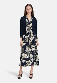 Marineblauwe bloemenjurk met witte en groene patronen, voorzien van een geknoopte taille en een lichte zwarte cardigan. Peep-toe schoenen maken de look compleet.