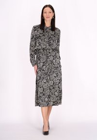 Czarna sukienka w wzór paisley z długimi rękawami, wysokim kołnierzem i zwężoną talią. Gładki materiał z płynącą spódnicą do kolan. Czarne szpilki.