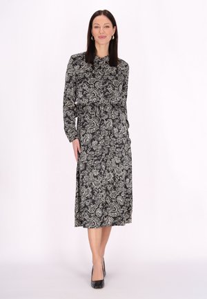 Sort kjole med paisley-mønster og lange ærmer, høj krave og indsnævret talje. Glat stof med en svævende, knælang nederdel. Sorte høje hæle.