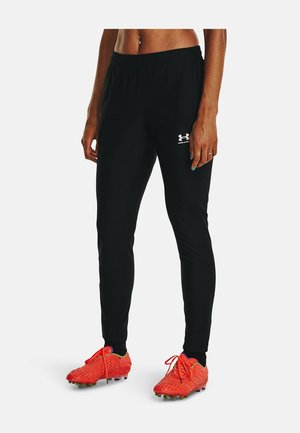 Personne portant un pantalon de sport noir Under Armour et des crampons orange vif, debout sur un fond blanc.