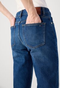 Persona che indossa jeans di denim blu con le mani infilarle nelle tasche posteriori, mostrando il logo del marchio e il patch in pelle sulla vita.