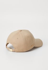 Beige baseball cap med buet skjerm, teksturert stoff og en gullfarget logosmykke på siden. Justerbar stropp bak.