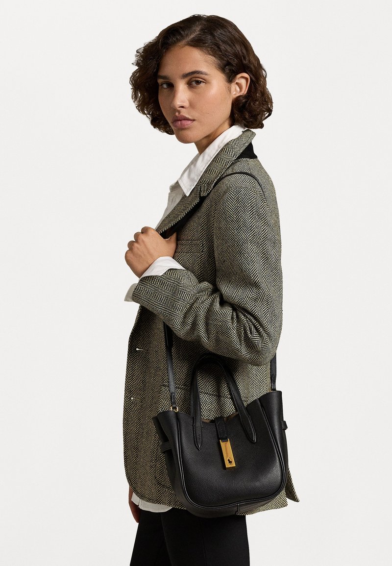 Borsa nera in pelle con una forma strutturata, manici superiori, tracolla regolabile e dettagli in oro. Indossata con un blazer a spina di pesce.