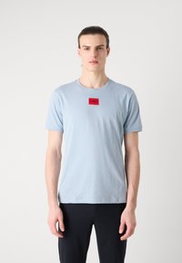Tricou din bumbac de culoare albastru deschis, cu mâneci scurte, guler rotund și un eticheta rectangulară roșie cu logo-ul "HUGO" pe piept.