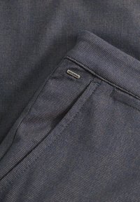 Tavă de material textil de culoare albastru închis, cu un model subtil, detaliu de buzunar și un mic tag metalic "GABBA" pe talie.