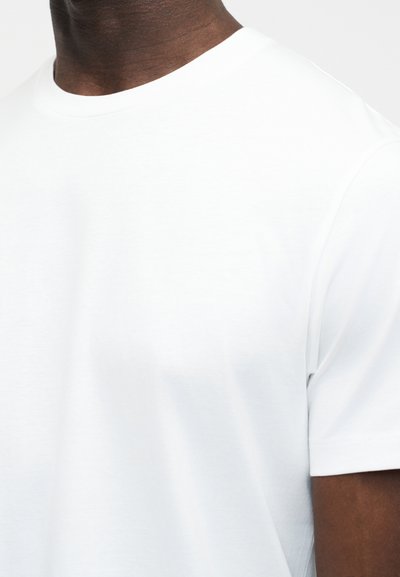 Άτομο με σκούρο δέρμα, που φοράει απλό λευκό κοντομάνικο μπλουζάκι με στρογγυλή λαιμόκοψη, κομμένο ώστε να φαίνεται ο λαιμός και το πάνω μέρος του στήθους.