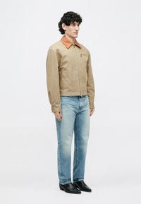 Homme debout portant une veste beige avec un col marron, un jean bleu clair et des bottes noires à bout pointu, sur un fond blanc uni.