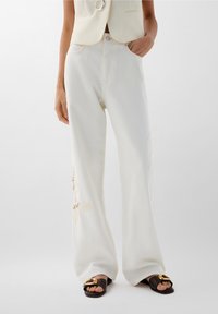 Pantaloni bianchi a gamba larga realizzati in tessuto morbido, con dettagli in pizzo sui pantaloni inferiori. Abbinati a sandali marroni con un accento dorato.