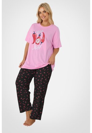 Mujer sonriente con camiseta rosa extragrande con la inscripción "Hot Stuff" y estampado de chile, combinada con pantalones negros con patrones rojos de chile, de pie descalza.