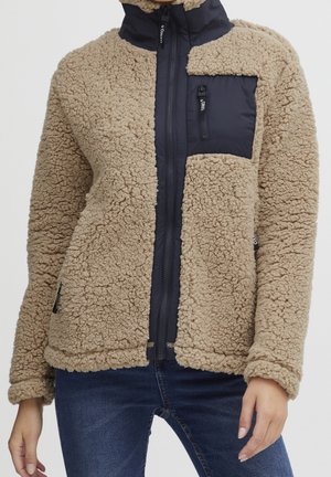 Veste en sherpa polaire beige avec une poche poitrine zippée bleu marine et une fermeture éclair frontale, portée avec un jean bleu.