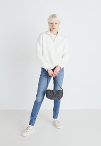 Valge tõmbetõmbluku ja tekstuuriga mustriga pullover, sinised teksad ja väike hall kott, millel on nupud, koos valgete tossudega.