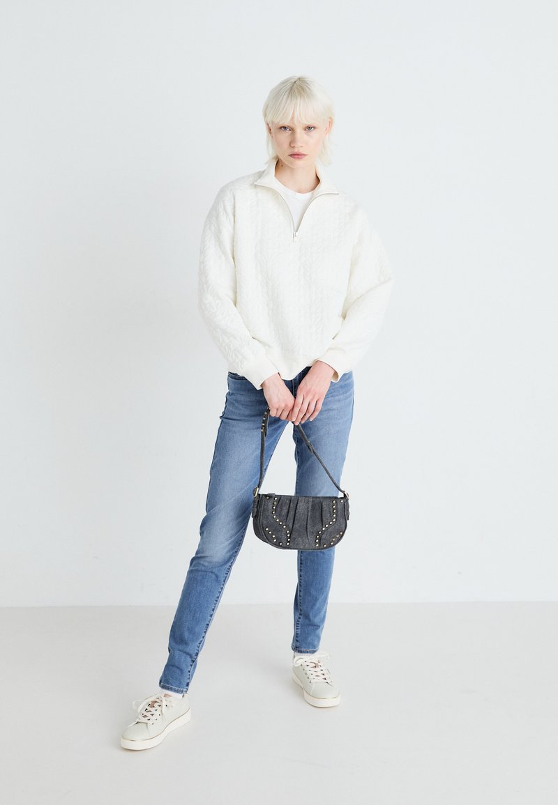Valge tõmbetõmbluku ja tekstuuriga mustriga pullover, sinised teksad ja väike hall kott, millel on nupud, koos valgete tossudega.