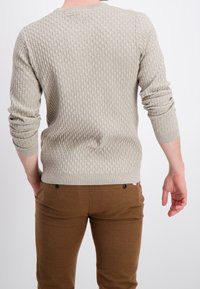 Světle béžový pletený svetr s texturou a vzorovaným povrchem, doplněný o hnědé přiléhavé kalhoty se dvěma knoflíky.