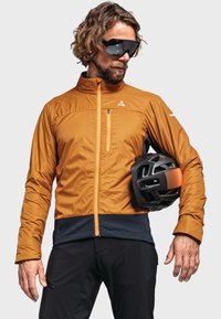 Veste isolante orange avec un col haut, section inférieure noire et fermeture éclair orange. Le modèle tient un casque noir avec des accents orange.