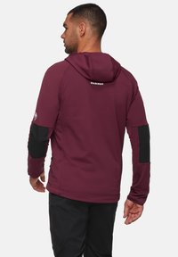 Veste à capuche bordeaux avec des accents noirs, tissu texturé et logo au dos. Avec des manches ajustées et des poches latérales zippées.