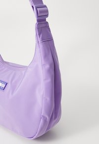 Violetti nailonista valmistettu olkalaukku, jossa on sileä pinta, pyöreä muoto ja säädettävä hihna, sekä pieni logo-tagi kontrastia edustavassa sinisessä.