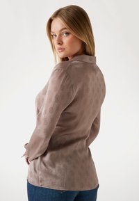 Pitkähihaista paitaa pehmeässä taupe-kankaassa, jossa on hillitty kohokuviointi, napilliset hihansuut ja kaareva helma.