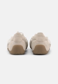 Ζευγάρι μπεζ σουέντ αθλητικά παπούτσια με καουτσούκ σόλες, εμφανιζόμενα από πίσω σε λευκό φόντο, με ανυψωμένο σχεδιασμό στη φτέρνα.