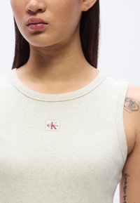 Ανοιχτό γκρι αμάνικο τοπ κατασκευασμένο από ριμ π ύφασμα, με μικρό λογότυπο Calvin Klein σε κόκκινο πάνω σε λευκό φόντο.