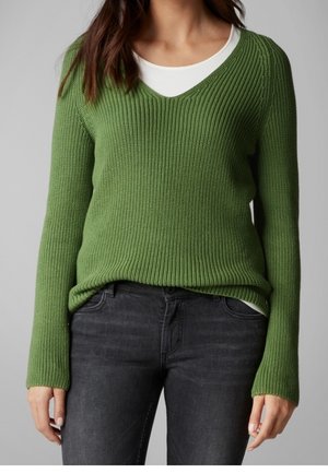 Femme portant un pull vert côtelé à col en V par-dessus un top blanc et un jean gris foncé, debout devant un fond gris.