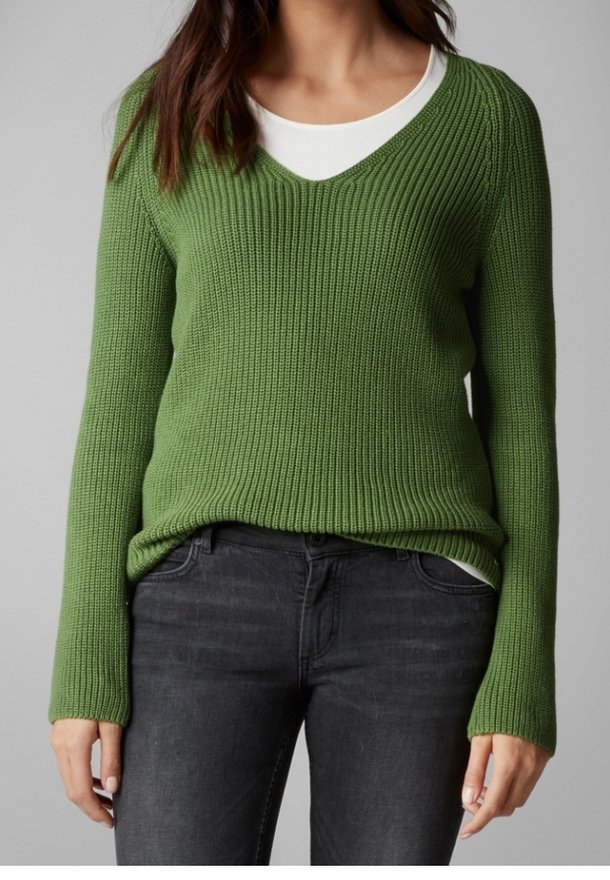 Femme portant un pull vert côtelé à col en V par-dessus un top blanc et un jean gris foncé, debout devant un fond gris.