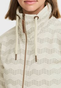 Veste zippée beige clair avec des motifs en zigzag, cordons de réglage crème et quincaillerie de fermeture éclair en bronze contrastante. Texture de tissu douce.