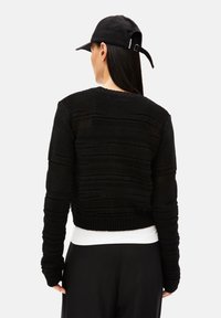 Schwarzer gestrickter, verkürzter Pullover mit strukturierten vertikalen Streifen, gerippten Bündchen und Saum, über einem weißen Oberteil getragen, mit einer sichtbaren schwarzen Mütze.