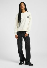 Sudadera crema con un diseño de corazón en el lado izquierdo del pecho, combinada con jeans negros de pierna ancha y zapatillas negras. Textura suave, corte relajado.