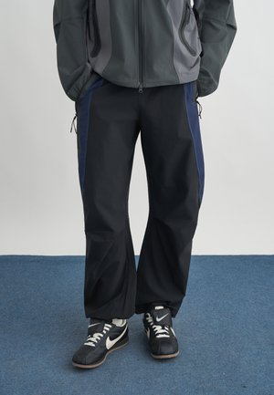 Άτομο που φοράει μαύρο και ναυτικό μπλε φόρμα, γκρι μπουφάν και μαύρα αθλητικά παπούτσια Nike, στέκεται σε μπλε μοκέτα.