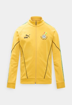 Κίτρινο αθλητικό μπουφάν με φερμουάρ, με το λογότυπο της Ποδοσφαιρικής Ομοσπονδίας της Γκάνας, το λογότυπο Puma, το κείμενο «KING» και πράσινες ριγέ μοτίβες στα μανίκια.