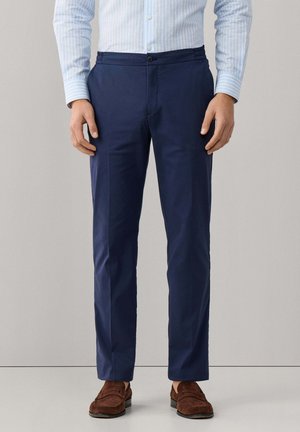 Homme portant un pantalon habillé bleu marine, une chemise rayée bleu clair et des mocassins en daim marron, debout devant un fond neutre.