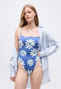 Γυναίκα που φοράει ολόσωμο μαγιό μπλε με μεγάλα λευκά μαργαρίτες και μια υπερμεγέθη ανοιχτό μπλε πουκάμισο κρεμασμένο στους ώμους της.