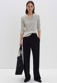 Donna con una camicia a maniche lunghe a quadri e pantaloni neri che tiene una borsa a tracolla nera, in piedi davanti a uno sfondo chiaro e uniforme.