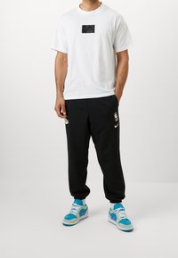 Bílá bavlněná tričko s černou nášivkou loga Nike, doplněné černými tepláky se logem NBA a modro-šedými teniskami s fialovými akcenty.