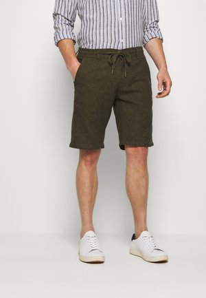 Homme portant un short vert olive à cordon, des baskets blanches et une chemise rayée à manches longues retroussée, debout avec une main dans la poche.
